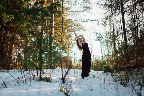 Portrait of a girl in a long dark dress, which stands in the winter Sunny forest among the trees. The girl stands in the pose of a tree, her hands are like branches, and her body is the trunk of a tree.