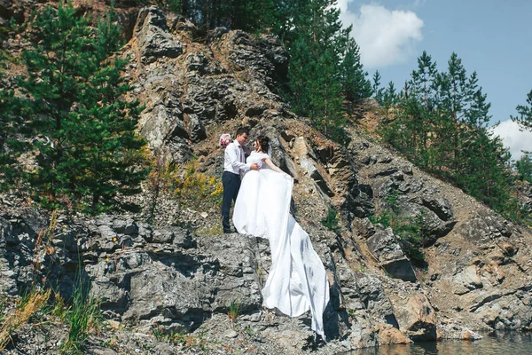 Felices recién casados parados en las rocas cerca del lago azul claro —  Fotos de Stock