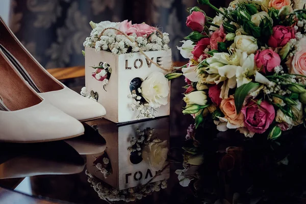 Lindas flores de casamento em uma cesta de madeira e sapatos de noiva branca em uma mesa de vidro escuro — Fotografia de Stock