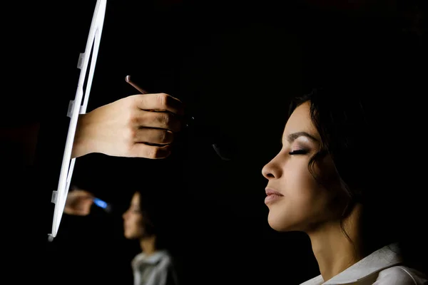 Makeup artist puts makeup on a beautiful brunette girl on a dark background. The concept of beauty