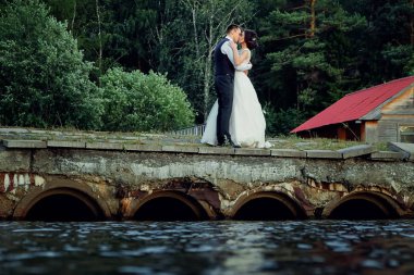 Şık bir beyaz elbise ve damat gelin eski bir taş köprü üzerinde stand, arkasında kalın bir yeşil orman