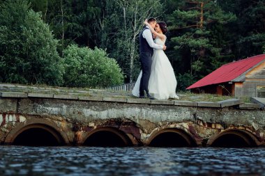 Şık bir beyaz elbise ve damat gelin eski bir taş köprü üzerinde stand, arkasında kalın bir yeşil orman