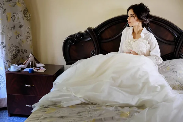 Bella piccola sposa triste seduta sul letto che abbraccia il suo vestito bianco il giorno del suo matrimonio — Foto Stock