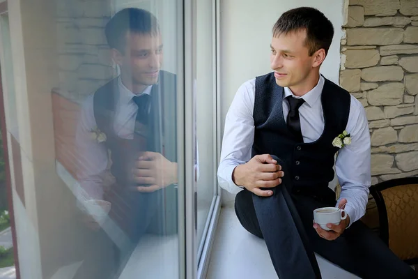 Ein schöner Mann sitzt auf der Fensterbank eines großen Fensters und trinkt seinen Morgenkaffee — Stockfoto