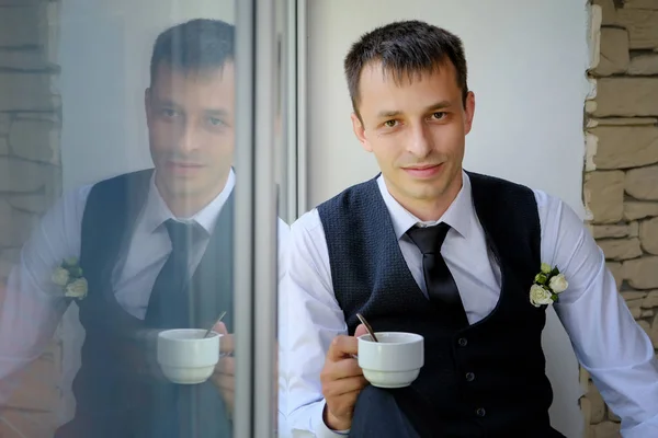 Un hombre guapo se sienta en el alféizar de una ventana grande y bebe su café de la mañana — Foto de Stock