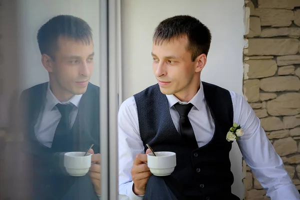 Un hombre guapo se sienta en el alféizar de una ventana grande y bebe su café de la mañana — Foto de Stock