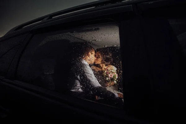 The bride and groom kissing in the car late at night, and it\'s raining outside. Bride and groom photographed behind the wet glass