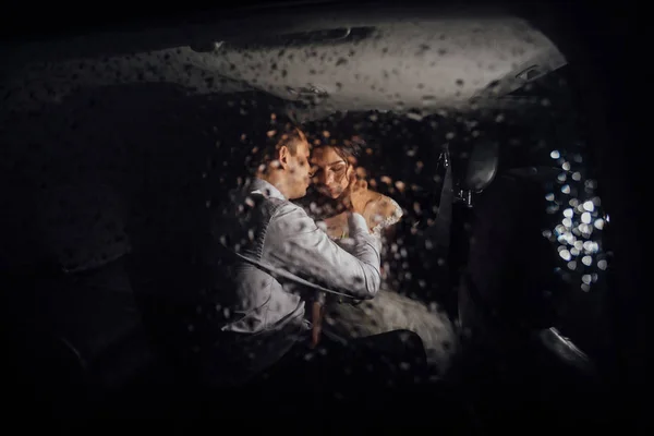 A noiva e o noivo a beijarem-se no carro à noite, e está a chover lá fora. Noiva e noivo fotografado atrás do vidro molhado — Fotografia de Stock