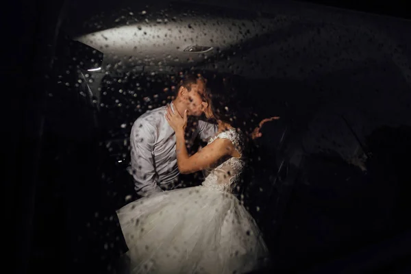 The bride and groom kissing in the car late at night, and it\'s raining outside. Bride and groom photographed behind the wet glass