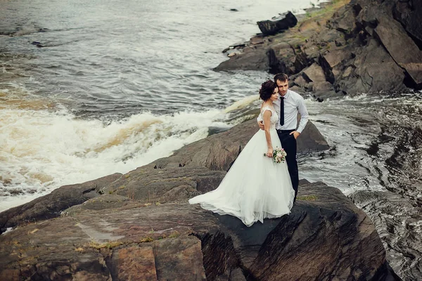 Η νύφη σε ένα πολυτελές λευκό φόρεμα και ένα όμορφο γαμπρός που αγκαλιάζει το υπόβαθρο της φύσης. Νεόνυμφοι είναι σε μια βραχώδη ακτή, γύρω από τους το νερό βράζει — Φωτογραφία Αρχείου