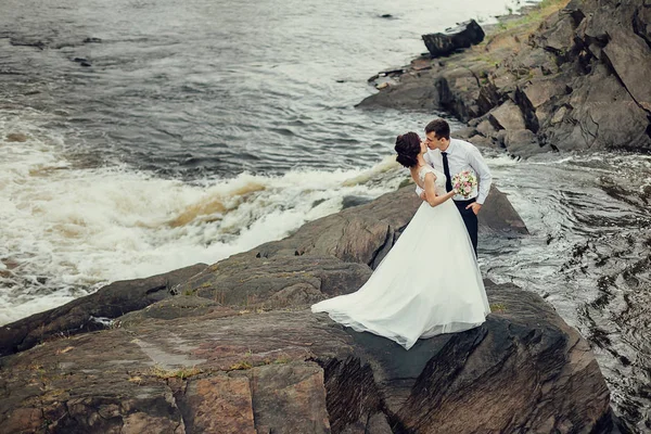 La novia en un lujoso vestido blanco y un hermoso novio abrazándose contra el fondo de la naturaleza. Los recién casados están en una costa rocosa, alrededor de ellos el agua hierve —  Fotos de Stock