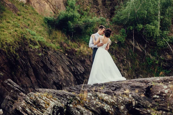 La novia en un lujoso vestido blanco y un hermoso novio abrazándose contra el fondo de la naturaleza. Los recién casados están en una costa rocosa, alrededor de ellos el agua hierve —  Fotos de Stock