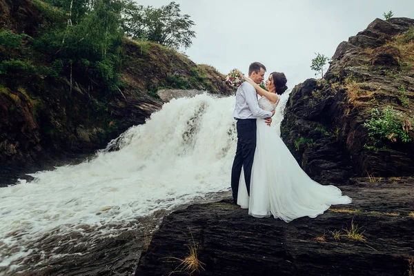 Happy bride and groom stand on the background of the waterfall and hugging — Stock Photo, Image