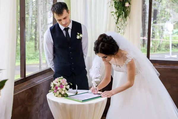 Braut und Bräutigam unterzeichnen das Dokument über die Hochzeit in der Nähe des mit Blumen geschmückten Hochzeitsbogens — Stockfoto