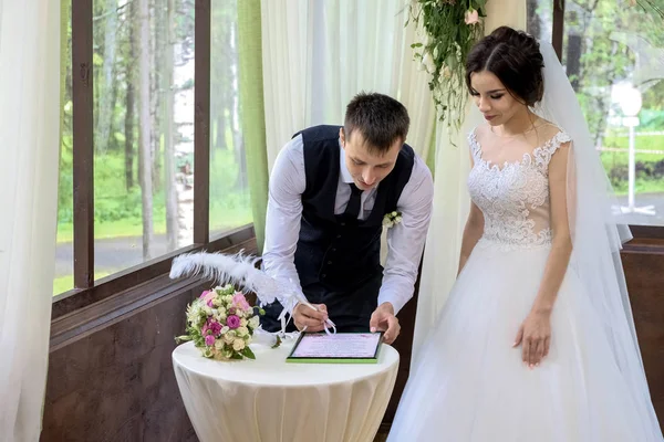 Braut und Bräutigam unterzeichnen das Dokument über die Hochzeit in der Nähe des mit Blumen geschmückten Hochzeitsbogens — Stockfoto