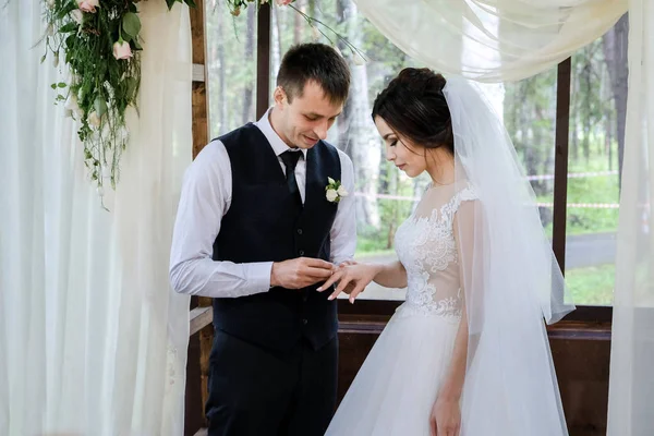 A noiva e noivo trocam anéis de casamento perto do arco de casamento decorado com flores — Fotografia de Stock