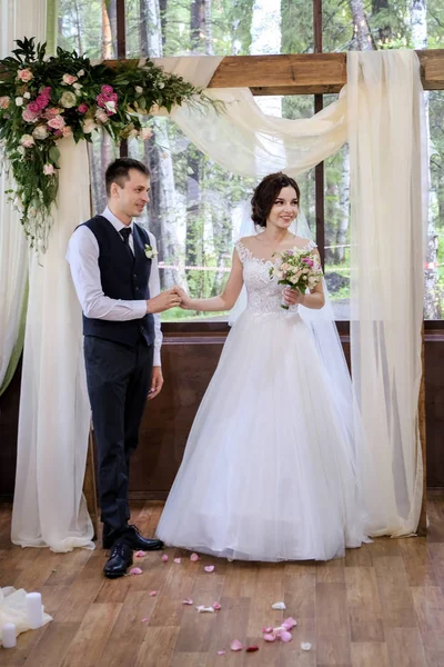 Bruden och brudgummen nära bröllop bågen dekorerad med blommor — Stockfoto