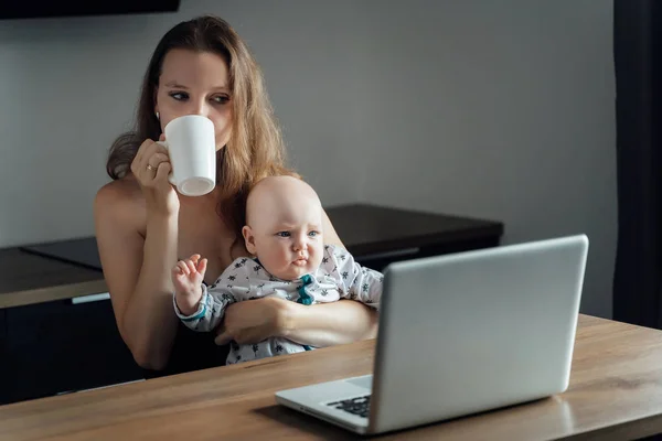 Young mother sits at a table with a baby in her arms and works o — Stock Photo, Image