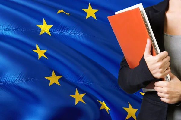 Learning European language concept. Young woman standing with the European Union flag in the background. Teacher holding books, orange blank book cover.