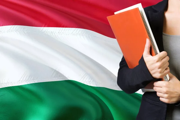 ハンガリー語の言語の概念を学習します バック グラウンドでハンガリーの国旗と立っている若い女性 先生の本 オレンジの空白ブック カバーを保持 — ストック写真
