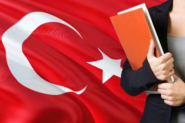 Learning Turkish language concept. Young woman standing with the Turkey flag in the background. Teacher holding books, orange blank book cover.