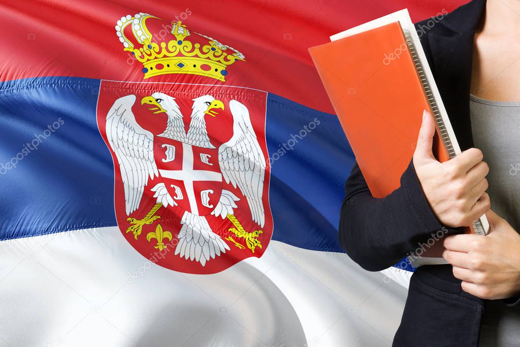 Learning Serbian language concept. Young woman standing with the Serbia flag in the background. Teacher holding books, orange blank book cover.