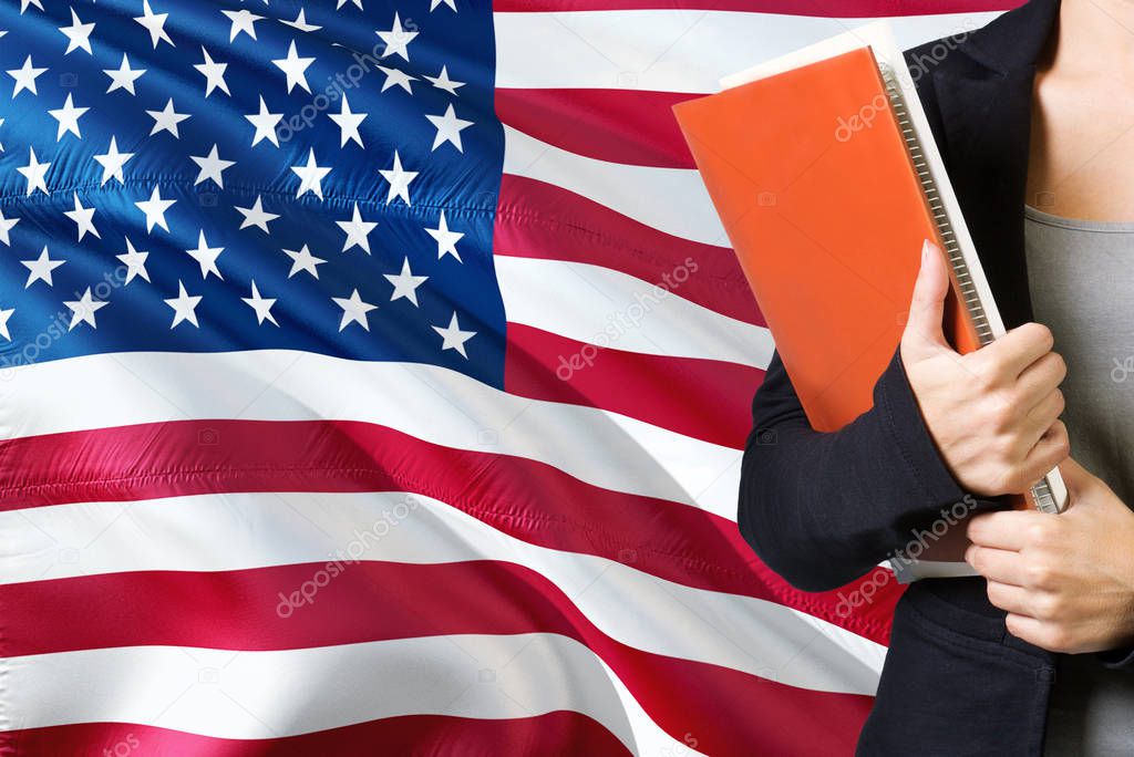 Learning American language concept. Young woman standing with the United States flag in the background. Teacher holding books, orange blank book cover.