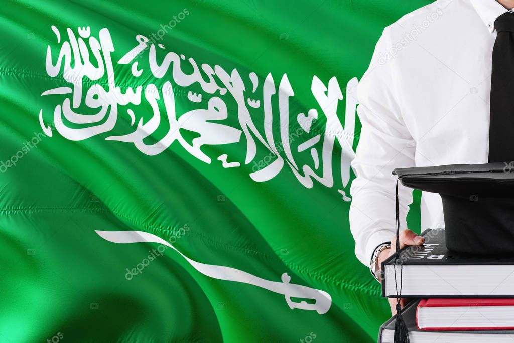 Successful Saudi student education concept. Holding books and graduation cap over Saudi Arabia flag background.