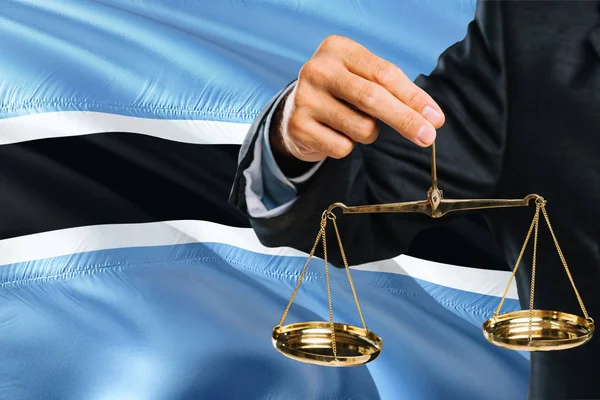 Judge is holding golden scales of justice with Botswana waving flag background. Equality theme and legal concept.