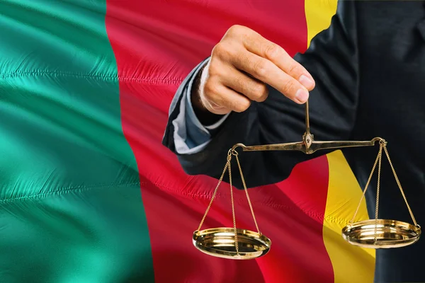 Cameroonian Judge is holding golden scales of justice with Cameroon waving flag background. Equality theme and legal concept.