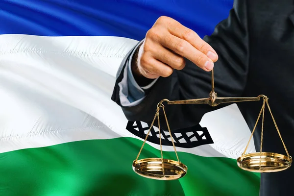 Judge is holding golden scales of justice with Lesotho waving flag background. Equality theme and legal concept.