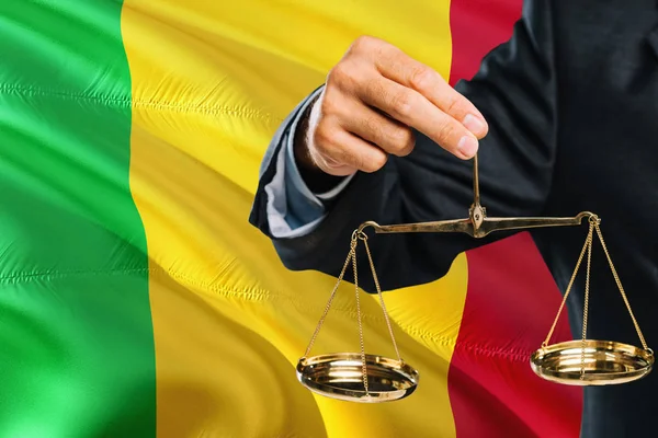 Malian Judge is holding golden scales of justice with Mali waving flag background. Equality theme and legal concept.