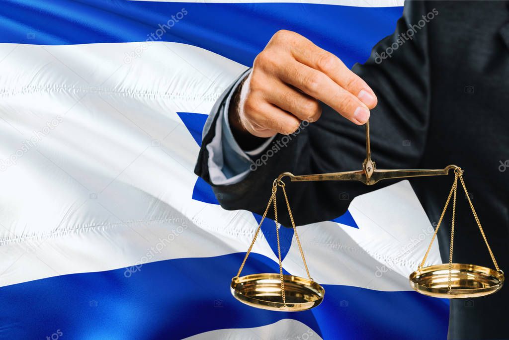 Israeli Judge is holding golden scales of justice with Israel waving flag background. Equality theme and legal concept.