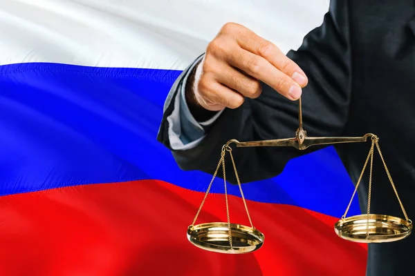Russian Judge is holding golden scales of justice with Russia waving flag background. Equality theme and legal concept.