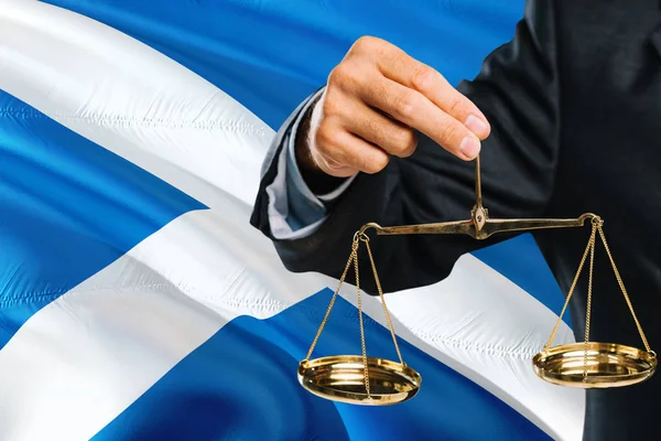 Scottish Judge is holding golden scales of justice with Scotland waving flag background. Equality theme and legal concept.