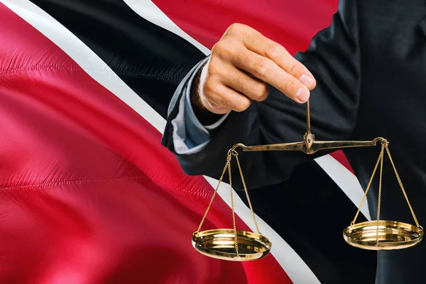 Judge is holding golden scales of justice with Trinidad And Tobago waving flag background. Equality theme and legal concept.