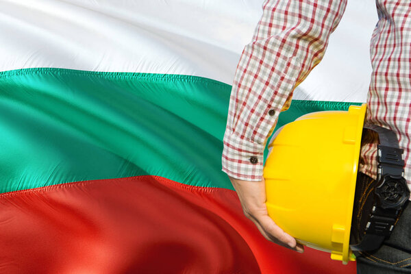 Bulgarian Engineer is holding yellow safety helmet with waving Bulgaria flag background. Construction and building concept.