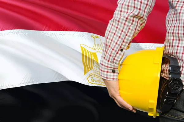 Egyptian Engineer is holding yellow safety helmet with waving Egypt flag background. Construction and building concept.