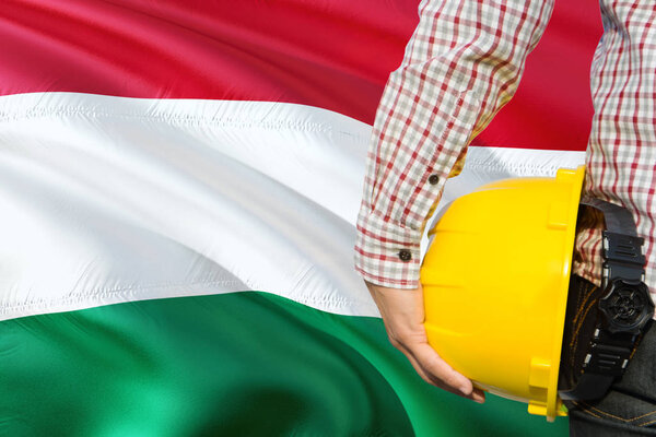 Hungarian Engineer is holding yellow safety helmet with waving Hungary flag background. Construction and building concept.