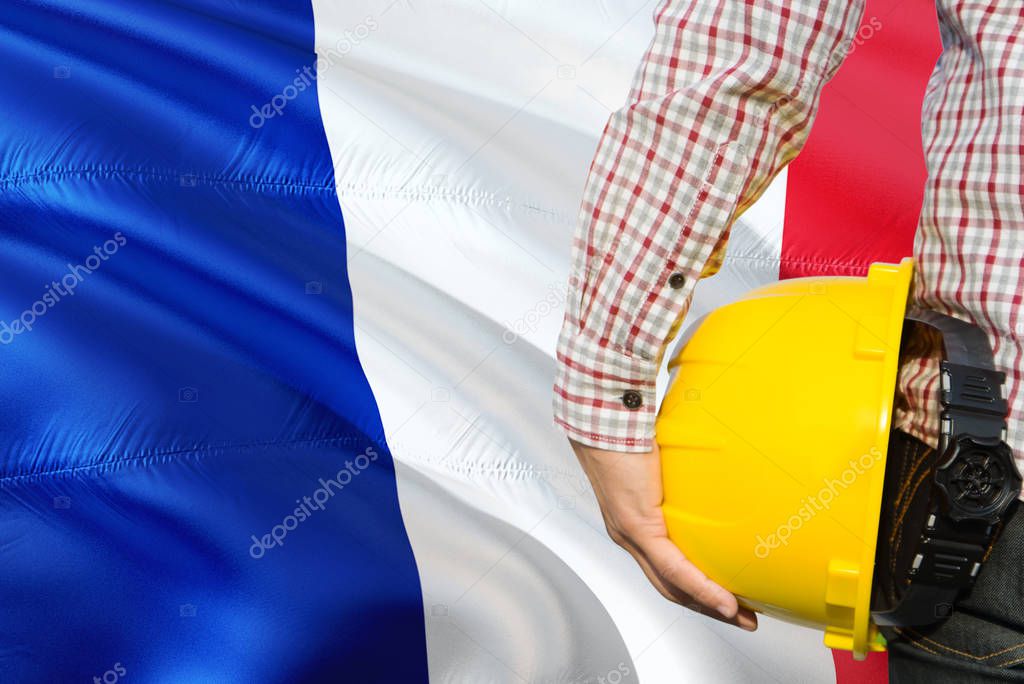 French Engineer is holding yellow safety helmet with waving France flag background. Construction and building concept.