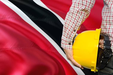 Mühendis sarı güvenlik kask sallayarak Trinidad ve Tobago bayrak arka plan ile tutuyor. İnşaat ve yapı kavramı.