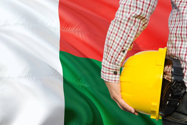 Malagasy Engineer is holding yellow safety helmet with waving Madagascar flag background. Construction and building concept.
