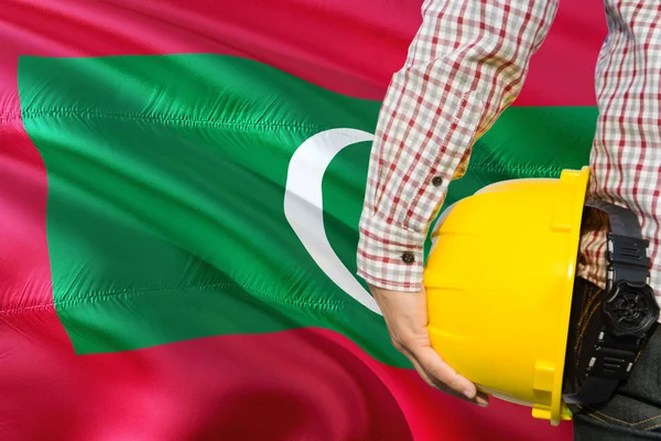 Maldivan Engineer is holding yellow safety helmet with waving Maldives flag background. Construction and building concept.