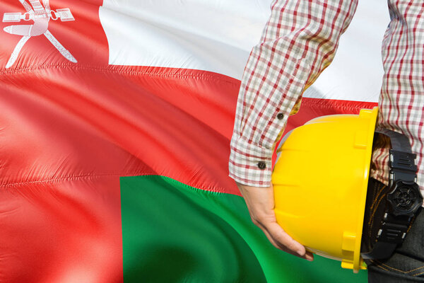 Omani Engineer is holding yellow safety helmet with waving Oman flag background. Construction and building concept.