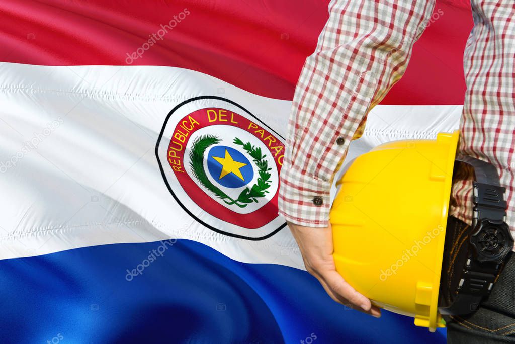 Paraguayan Engineer is holding yellow safety helmet with waving Paraguay flag background. Construction and building concept.