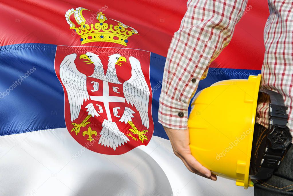 Serbian Engineer is holding yellow safety helmet with waving Serbia flag background. Construction and building concept.