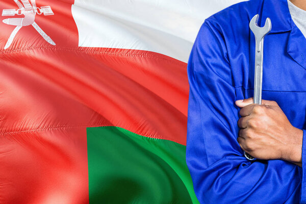 Omani Mechanic in blue uniform is holding wrench against waving Oman flag background. Crossed arms technician.