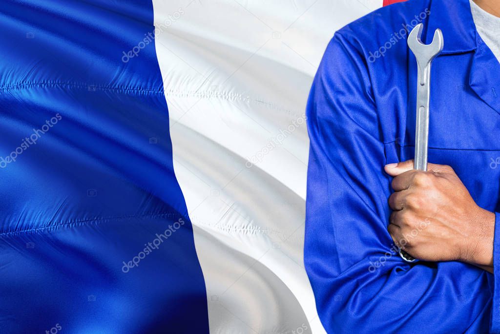 French Mechanic in blue uniform is holding wrench against waving France flag background. Crossed arms technician.