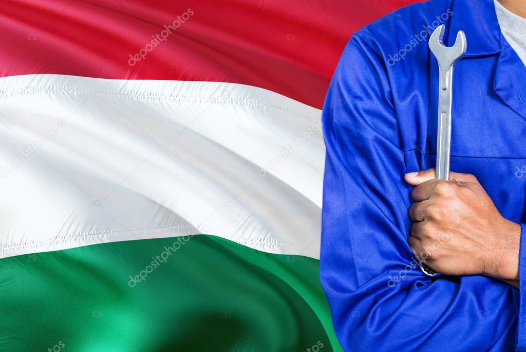 Hungarian Mechanic in blue uniform is holding wrench against waving Hungary flag background. Crossed arms technician.