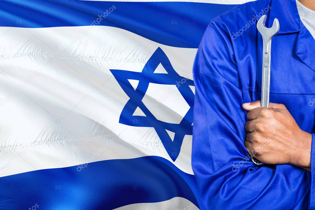 Israeli Mechanic in blue uniform is holding wrench against waving Israel flag background. Crossed arms technician.